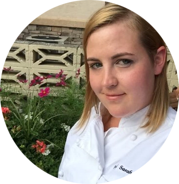 chef sarah stands in front of a bed of flowers. she is wearing a chef jacket and has blonde, shoulder length hair